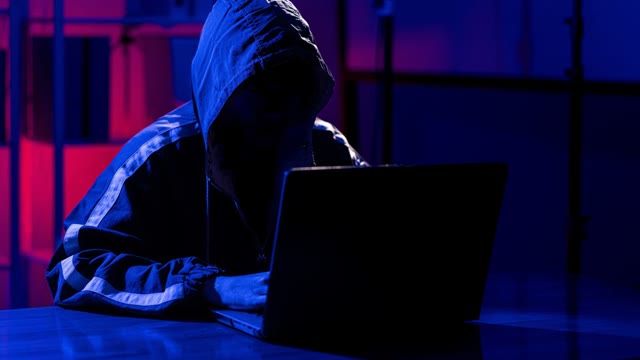 A hooded figure working at a laptop in a dark room